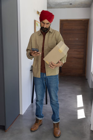 Indian man holding taped package and using smartphone by console table with framed art in entryway....