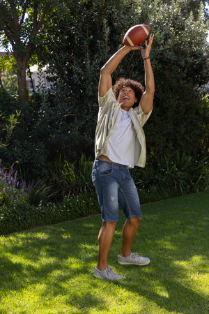 Man catching American football on backyard grass lawn under sunlight filtering through trees....
