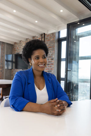 African American woman sitting at white conference table in office using monitor near city skyline....