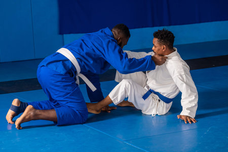 African American male training partners grappling on blue mats in dojo wearing gis and belts....