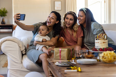 Diverse female friends holding baby boy taking selfie with smartphone on sofa with birthday cake....
