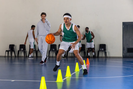 African American male basketball player dribbling orange basketball through yellow cones on court....