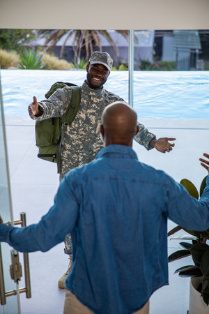 African American father and son embracing at home entrance in camouflage uniform carrying backpack....