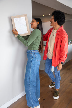 African American and Indian couple hanging wooden-framed picture on white wall in kitchen area....