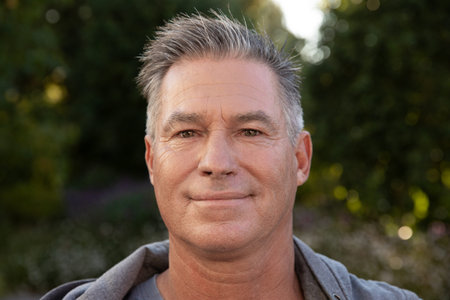 Middle-aged man standing calmly in backyard garden wearing grey hoodie under soft sunlight....