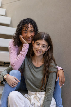 Female friends sitting on white staircase against beige wall wearing casual tops, smiling at camera....