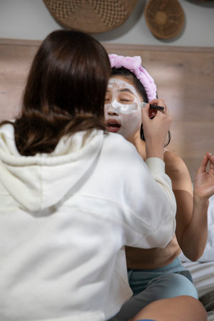 Diverse women friends applying facial mask with brush on bed with pink headband by woven baskets....