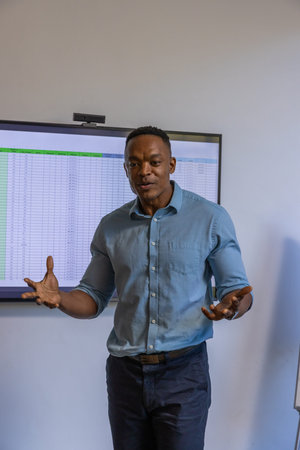 African American man gesturing, speaking in meeting room with screen showing spreadsheet and webcam....