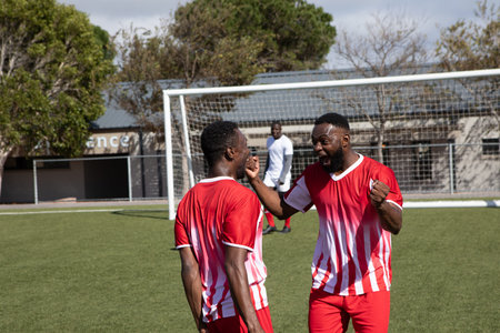 African American male teammates celebrating goal in red-and-white kits on soccer field near net....