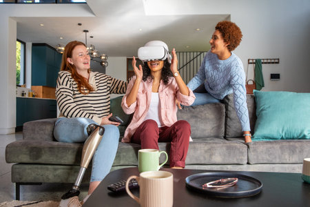 Diverse female friends sitting on sofa in living room adjusting VR headset with prosthetic leg. Technology, socializing, modern, cozy, casual, lifestyle, innovationの写真素材