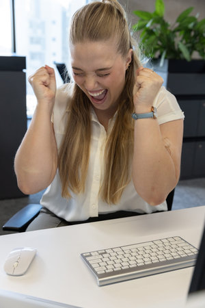 Celebrating with fists raised, woman in white blouse at office desk with white keyboard and mouse. Celebration, joy, workspace, modern, corporate, cityscape, daylightの写真素材