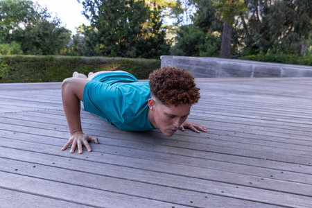 Athletic male performing push-ups in turquoise sportswear on wooden deck beside bench and hedges....