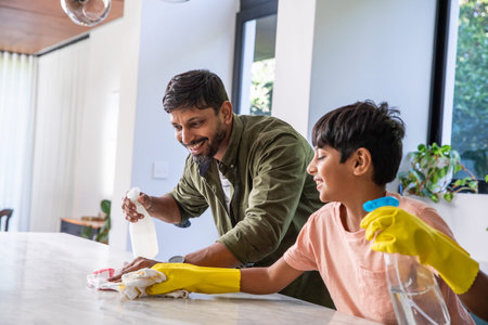 South Asian father and son cleaning marble kitchen island at home with spray bottles and cloths....