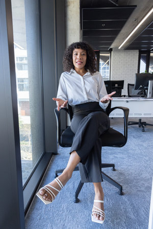 African American woman wearing work attire sitting at desk using keyboard with monitors near window....