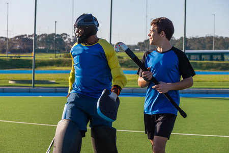 Male field hockey teammates standing on pitch near fence wearing goalie helmet holding hockey stick....
