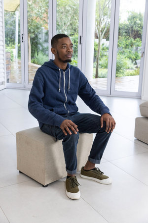 African American man in twenties sitting on beige cube ottoman in living area near glass doors....