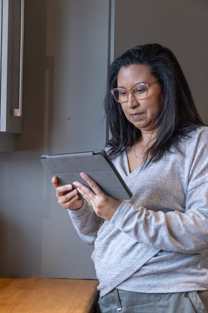 Woman standing beside wooden countertop beneath grey wall cabinet in home kitchen area using tablet....