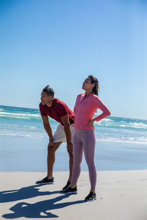 Diverse couple catching breath and stretching on sandy beach at ocean edge wearing running shoes....
