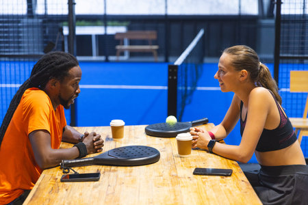 Diverse friends sitting at table in padel court lounge holding rackets with padel ball between them....