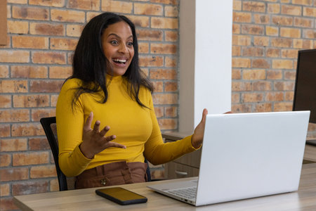 African American woman raising hands cheering project win at desk using laptop and phone....