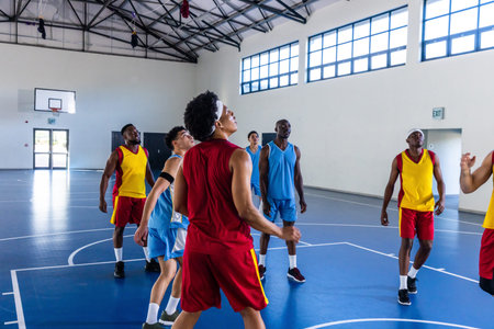 Diverse male basketball players reaching for rebound on bright blue court in gymnasium under hoop....