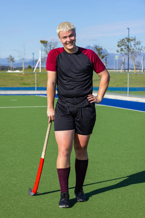 Male athlete smiling while gripping field hockey stick on synthetic turf with narrow water channel. Sport, outdoor, athletic, recreational, vibrant, energetic, competitionの写真素材