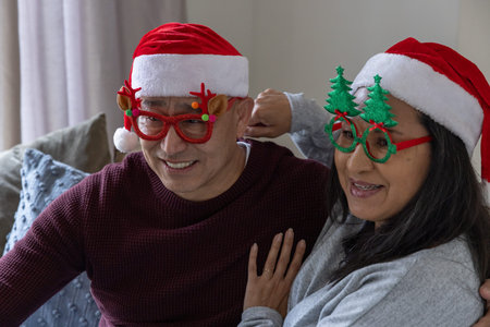 Senior couple sitting on sofa at home wearing Santa hats and novelty glasses among cushions....