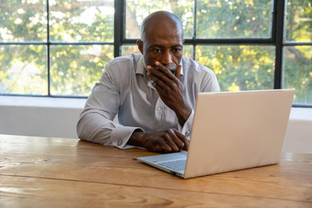 African American man wearing grey shirt sitting at wooden table working on laptop in office....
