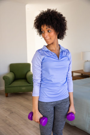 African American woman lifting purple dumbbells in home workout near green armchair and table lamp....