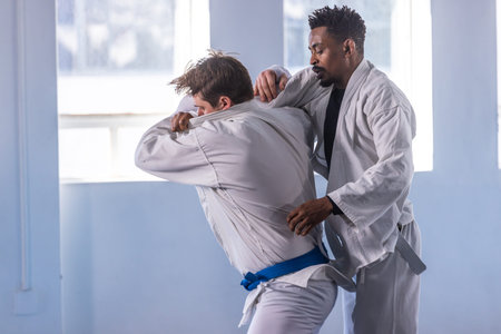 Diverse male martial artists grappling in training hall wearing white gis with grey and blue belts....