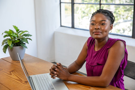 African American woman sitting in home office at wooden desk using laptop near potted houseplant....