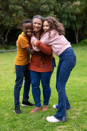 African American grandmother with teen grandchildren hugging on backyard lawn with flowers. Family,...