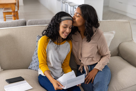 Mother kissing teenage daughter's forehead while reading letter on beige sofa with smartphone....