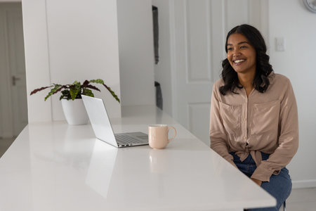 Woman sitting at kitchen island using laptop next to pink mug and planter, copy space. Minimalist,...
