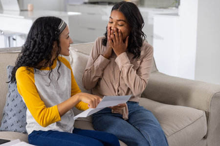 Diverse sisters sitting on light-gray sofa by kitchen island holding printed document and reacting....