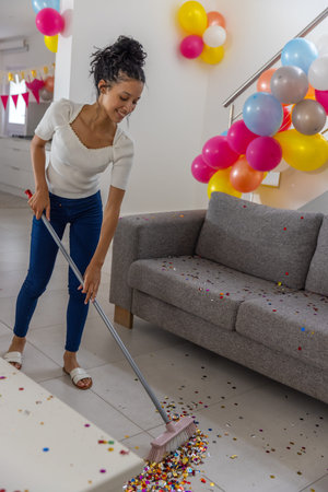 Teenage girl sweeping colorful confetti on living room floor with broom and balloons, copy space. Celebration, festivity, decor, vibrant, cheerful, leisure, cleanupの写真素材