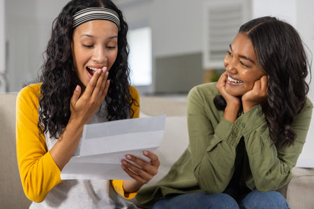 Diverse sisters sitting sofa reading letter and displaying surprise in living room. Families,...