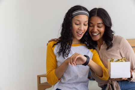 Diverse mother and teenage daughter opening gift box and examining smartwatch on bed in bedroom....