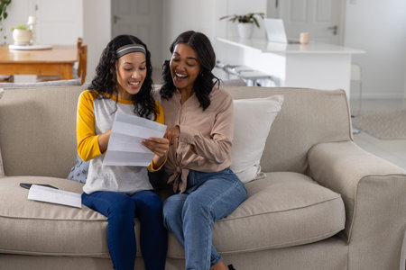 Diverse mother and teenage daughter sitting on sofa reading letter with envelope in living area. Family, bonding, communication, domestic, cozy, lifestyle, affectionの写真素材