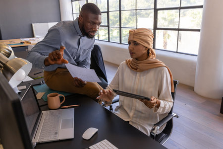 Diverse coworkers leaning on desk pointing at laptop and holding tablet at modern office space....