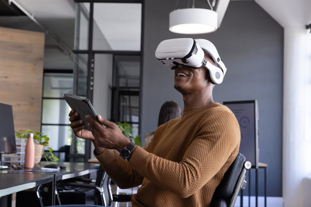 African American man using white VR headset and holding tablet at desk in open-plan office....