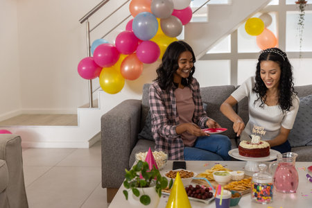 Diverse female friends slicing cake topped with 18 candle in living room amid balloons and snacks....