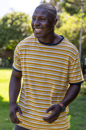African American male smiling and gesturing on sunlit backyard wearing striped T-shirt, wristwatch....