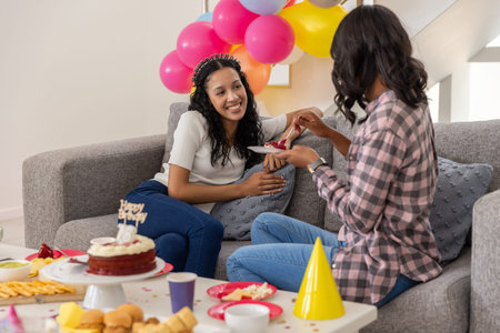 Diverse female friends sharing birthday cake on sofa in living room with balloons, copy space....