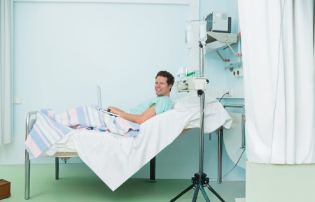 Man reclining on hospital bed in patient room, wearing gown, receiving IV infusion and using laptop....
