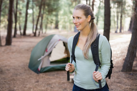 Female hiker wearing light green top standing in pine campsite holding backpack near green tent....