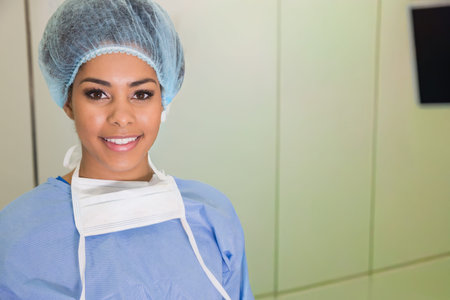 Adult female clinician standing looking at camera in OR wearing blue gown, bouffant cap, mask. Nurse, scrubs, medical, hospital, monitor, bright, sterileの写真素材