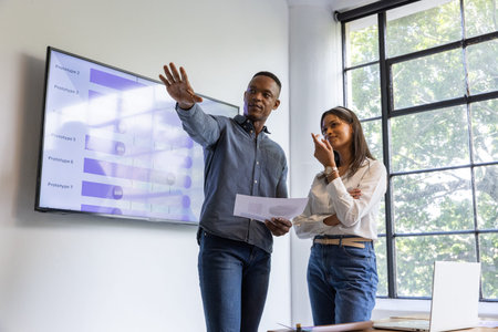 Diverse coworkers standing beside wall-mounted monitor reviewing printed charts in meeting room....