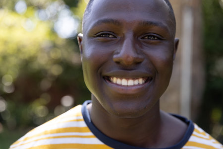 African American man smiling and wearing yellow striped shirt standing in backyard with foliage....