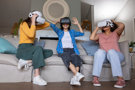 Diverse female friends wearing VR headsets and reacting on light gray couch in living room. Technology, innovation, entertainment, relaxation, modern, cozy, socialの写真素材